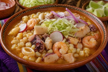 Plato de barro con caldo, maíz, camarones, pulpo, rábano, cebolla y lechuga, rodeado de recipientes con limones y rábanos.