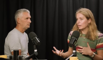 Un hombre y una mujer, Spector y Amati, sentados con micrófonos, conversan en un estudio. El hombre tiene cabello gris, la mujer rubio y lleva un suéter de rayas