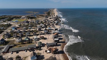 Vista aérea de una larga costa arenosa con casas elevadas y tejados oscuros que se extienden a lo largo de la playa, con el océano a la derecha y una laguna a la izquierda