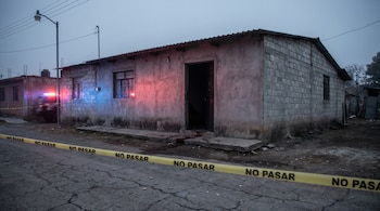 Fotografía de una vivienda humilde acordonada con cinta amarilla 'NO PASAR' en Tehuitzingo, Puebla. Luces de patrulla iluminan la fachada, casquillos en el suelo y manchas oscuras en la entrada.