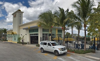 Exterior de un restaurante con una torre, en tonos claros y verdes, con varias palmeras, un SUV blanco estacionado y una calle en primer plano
