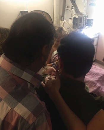 Primer plano de la cabeza de un bebé con cabello claro y los rostros de dos adultos que lo observan en una cama de hospital con equipamiento médico