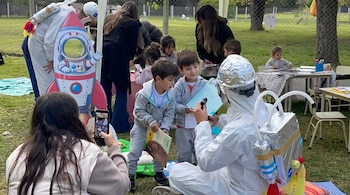Niños observan a una persona vestida de astronauta mientras una mujer fotografía la escena con un teléfono móvil en un parque.