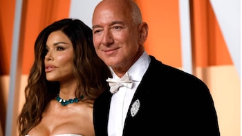 Jeff Bezos, um homem careca de smoking preto e gravata branca, sorri ao lado de Lauren Sánchez, de cabelos castanhos e colar turquesa, diante de um fundo laranja.