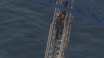 Persona con casco rojo y arnés escalando una estructura metálica tipo escalera sobre un cuerpo de agua oscuro y ligeramente agitado