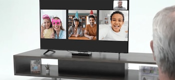 Un adulto mayor, de espaldas, observa una pantalla de televisión mostrando una videollamada con cinco personas sonrientes