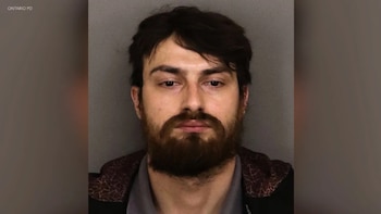 Fotografía de ficha policial de un hombre con barba, cabello oscuro y chaqueta sobre un fondo gris