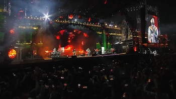 Escenario iluminado con músicos tocando instrumentos frente a una multitud de personas que sostienen teléfonos móviles encendidos