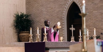 Un sacerdote con sotana morada levanta una hostia sobre un cáliz dorado en un altar con velas encendidas, un crucifijo y una planta