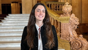 Mujer joven con cabello largo castaño, camisa blanca y chaqueta negra sonriendo; detrás, una escalinata de mármol con barandal tallado y ornamentado