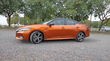 Un automóvil sedán de color naranja metálico con techo negro estacionado sobre grava junto a una hilera de árboles