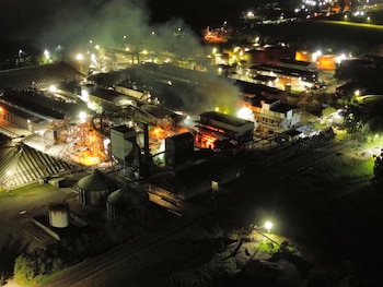 Planta industrial de noche con un incendio activo, mostrando llamas anaranjadas y humo denso sobre varios edificios. Luces artificiales iluminan la escena y tanques