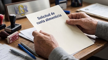 Primer plano de manos de adulto mayor sosteniendo una hoja con "Solicitud de cuota alimenticia", sobre un escritorio con sellos, bolígrafos y una insignia institucional borrosa.