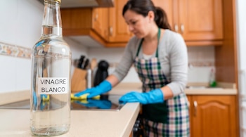 Botella de vinagre blanco con etiqueta en primer plano. Al fondo, una mujer con delantal y guantes azules limpia la encimera de una cocina.