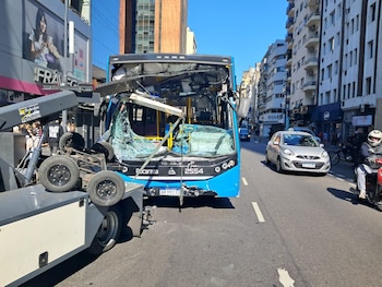 Un autobús azul con el parabrisas roto y la cabina dañada junto a un vehículo de remolque en una calle con automóviles