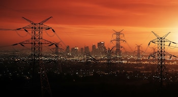 Varias torres de alta tensión y sus cables se erigen frente al horizonte de una ciudad con edificios y luces al anochecer bajo un cielo rojizo.