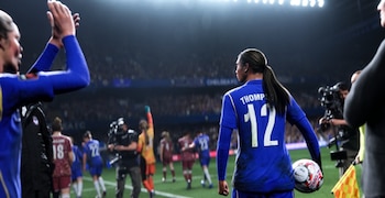Una futbolista vista de espaldas con camiseta azul número 12 lleva un balón en el campo de un estadio iluminado