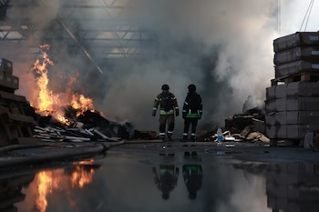Dos bomberos en uniforme caminan sobre el suelo mojado entre escombros humeantes, fuego e hileras de cajas apiladas