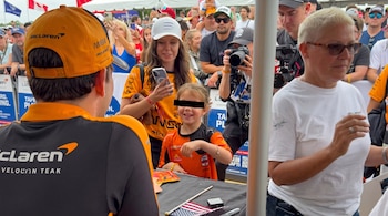Un piloto con uniforme de McLaren conversa con una niña sonriente frente a una multitud de espectadores y fotógrafos.