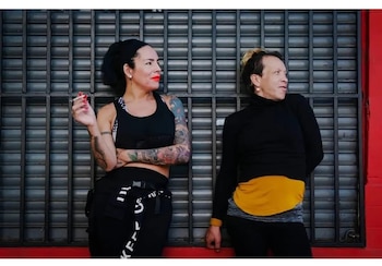 Dos personas, una con tatuajes y un cigarrillo, y otra con cuello alto, posan de pie frente a una persiana metálica gris con un marco rojo