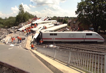 Vagones destrozados de un tren ICE blanco y rojo con personal de emergencia en chalecos y cascos naranjas. Vías de tren, una carretera elevada, casas y árboles