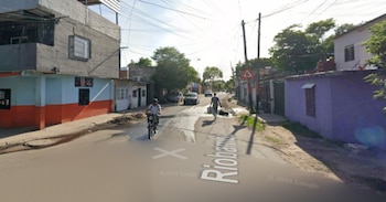 Una calle de barrio con casas, árboles y postes. Un motociclista y un peatón circulan. Automóviles estacionados. Señal de tránsito y texto "Riobamba" en el asfalto