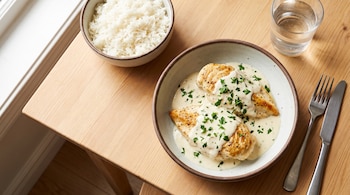 Dos pechugas de pollo con salsa blanca y perejil en un plato, un cuenco de arroz blanco, un vaso de agua, un tenedor y un cuchillo sobre una mesa de madera.