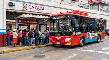 Un autobús rojo Binnibus está en una parada mientras un grupo de personas espera para abordar. Se ven letreros con 'Oaxaca' y 'Yuroo Viguera'.
