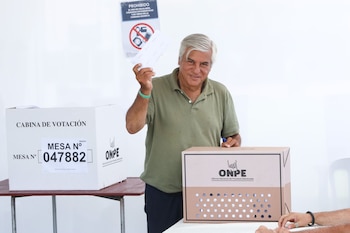 Fernando Olivera, un hombre de cabello gris y polo verde oliva, sonríe sosteniendo una papeleta en una cabina de votación de ONPE