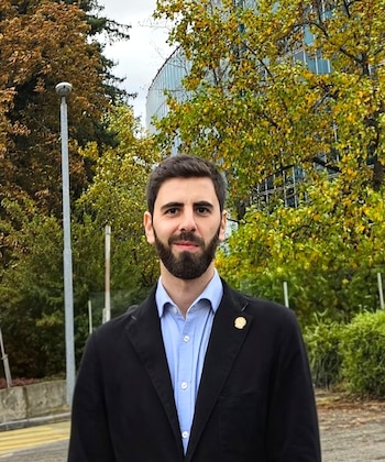 Un hombre con barba, camisa celeste y saco negro está de pie frente a árboles y un edificio con fachada de vidrio
