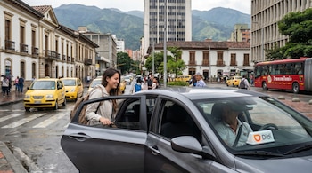 Una mujer sonriente entrando a un auto gris con un letrero Didi en el tablero, en una calle urbana de Bogotá con taxis amarillos, un bus TransMilenio y edificios.