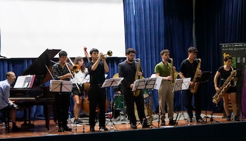 Siete músicos tocan en un escenario: pianista, trombonista, trompetista, contrabajista y tres saxofonistas. Fondo oscuro con cortinas azules