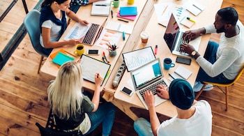 Cuatro personas trabajan en dos mesas de madera con laptops, documentos, gráficos, cuadernos, bolígrafos, lápices, tazas de café y teléfonos móviles.