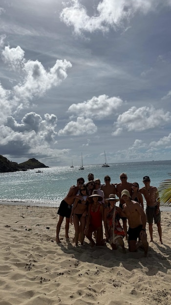 Grupo de aproximadamente doce personas en una playa de arena clara, posando frente al mar azul con barcos y montañas en la distancia bajo un cielo nublado