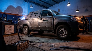 Una camioneta con placas de metal soldadas en las puertas dentro de un taller con herramientas en el suelo y chispas al fondo.