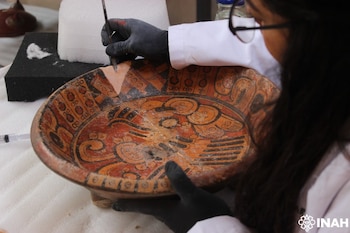 Manos con guantes negros usan un instrumento sobre el borde de un plato cerámico antiguo pintado con motivos prehispánicos