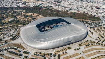 Campo de fútbol verde con arcos blancos dentro de un estadio con gradas repletas de aficionados. Montañas y cielo nublado se ven al fondo