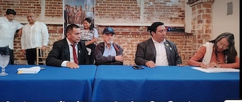 Cuatro personas sentadas en una mesa azul; una mujer firma un papel. Tres personas de pie detrás. Fondo de pared de ladrillo y un cartel con mujeres