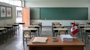 Aula escolar vacía con pupitres ordenados, una pizarra verde y un escritorio con carpetas, cuadernos y una pequeña bandera de Perú.