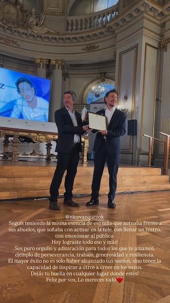 Dos hombres de traje en un escenario, uno entrega un diploma a otro. Detrás, una pantalla exhibe el rostro de un hombre. Edificio con columnas y adornos