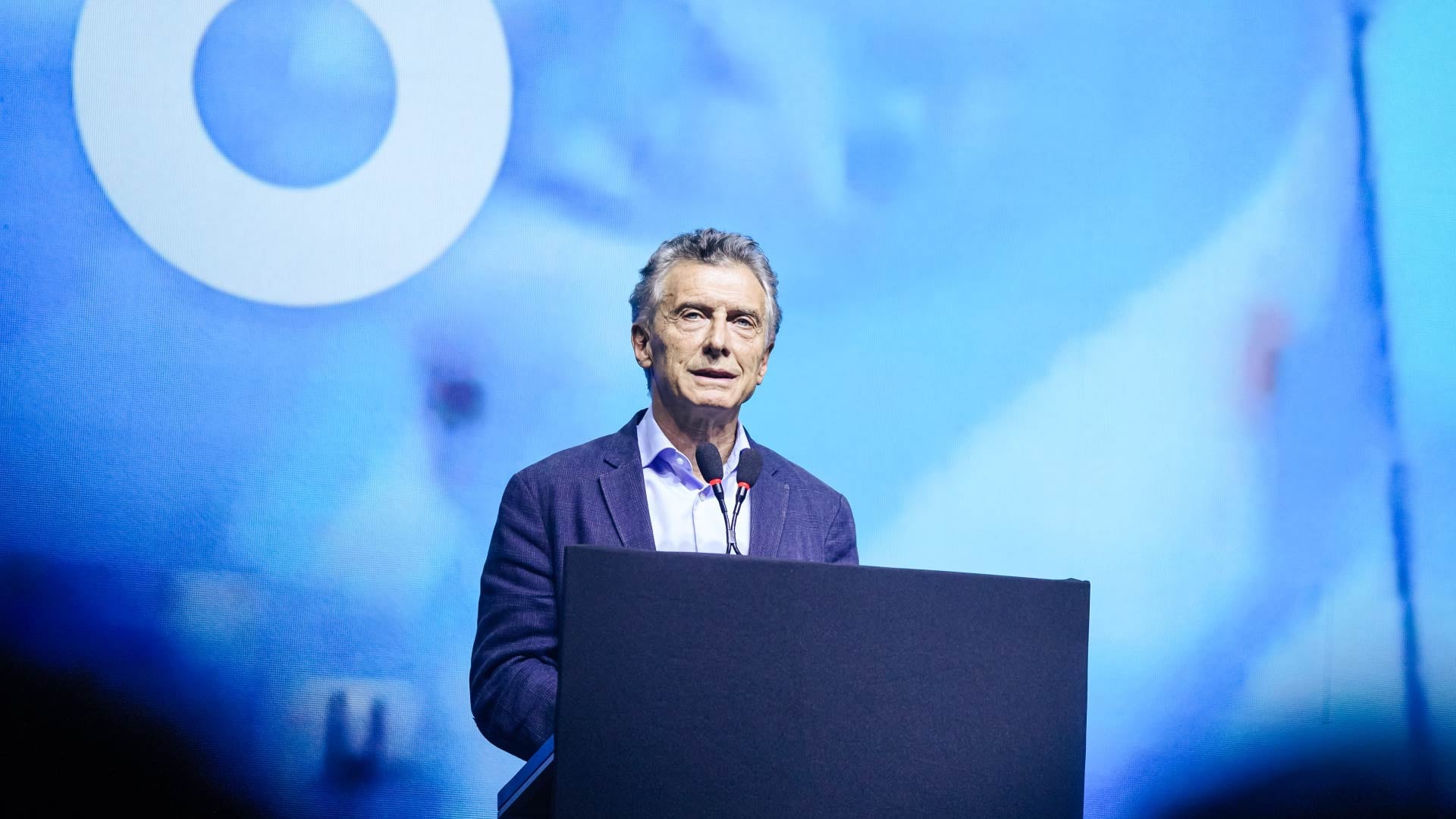 Mauricio Macri, expresidente de Argentina, pronuncia un discurso desde un atril, con un fondo azul que exhibe un logotipo parcial.