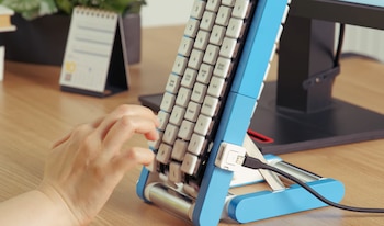 Mano sobre las teclas de un teclado blanco colocado en posición vertical en un soporte azul sobre un escritorio de madera
