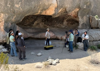 Grupo de nueve personas en un abrigo rocoso. Mujer en plataforma, cámara en trípode. Rocas grandes, marcas rojizas en pared. Suelo de tierra, vegetación dispersa
