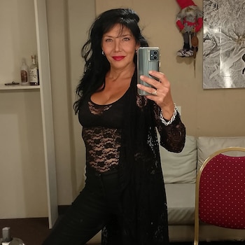 Una mujer de cabello oscuro y vestimenta negra sostiene un teléfono celular frente a un espejo en un ambiente interior