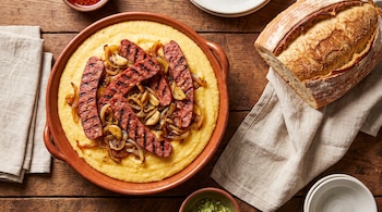 Fuente con polenta, tiras de salchicha parrillera, cebolla, ajo, pan casero, servilletas de tela y cuencos sobre mesa de madera.