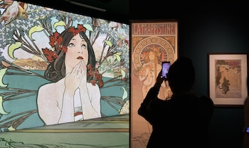 Una persona de espaldas fotografía con un teléfono móvil carteles ilustrados de estilo Art Nouveau expuestos en una sala