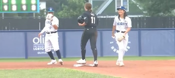 Tres jugadoras de béisbol en campo. Una con uniforme negro y casco, dos con uniforme a rayas, gorra azul y guantes. Marcador y valla azul de fondo