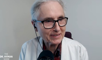 Primer plano de un hombre mayor con gafas y cabello gris, vestido con bata blanca, camisa a cuadros y auriculares in-ear, hablando frente a un micrófono