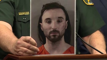 Un oficial de uniforme sostiene la fotografía de un hombre con barba en ropa naranja detrás de un atril con micrófono