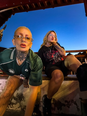 Dos mujeres rubias, una con cabello corto y otra largo, usan lentes y tienen múltiples tatuajes. Posan al aire libre bajo un cielo oscuro